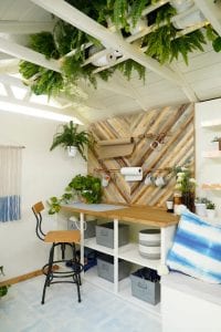 Shed detail: Wooden table top and tall chair. Plants and rolls of material on wall.
