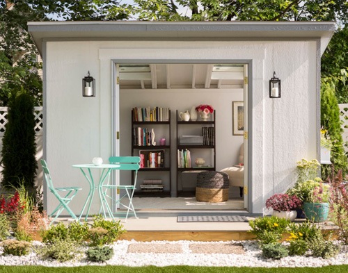 Indoor/Outdoor She Shed: Studio style white shed with doors open. Interior shows two book shelves and chair. Exterior shows green table and two chairs.